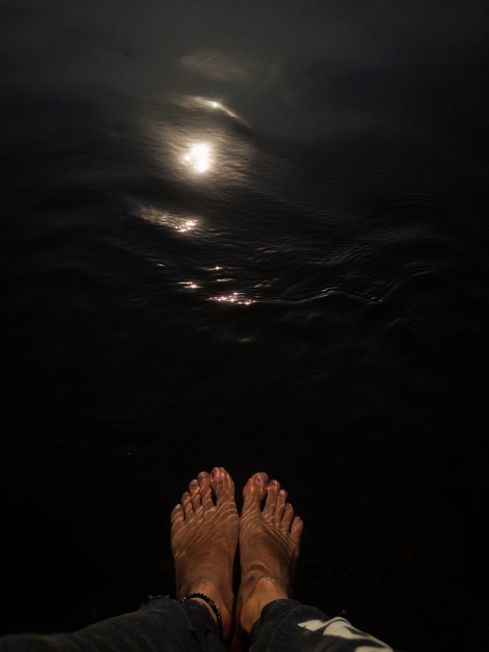 Feet by water reflecting the moonlight at night, creating a serene and calm atmosphere.