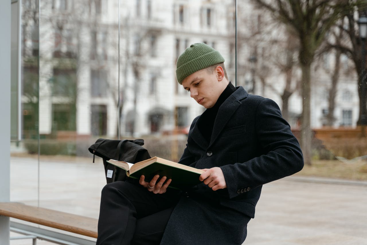 A young man in a black coat and green knitted cap reading a book outdoors in a park.