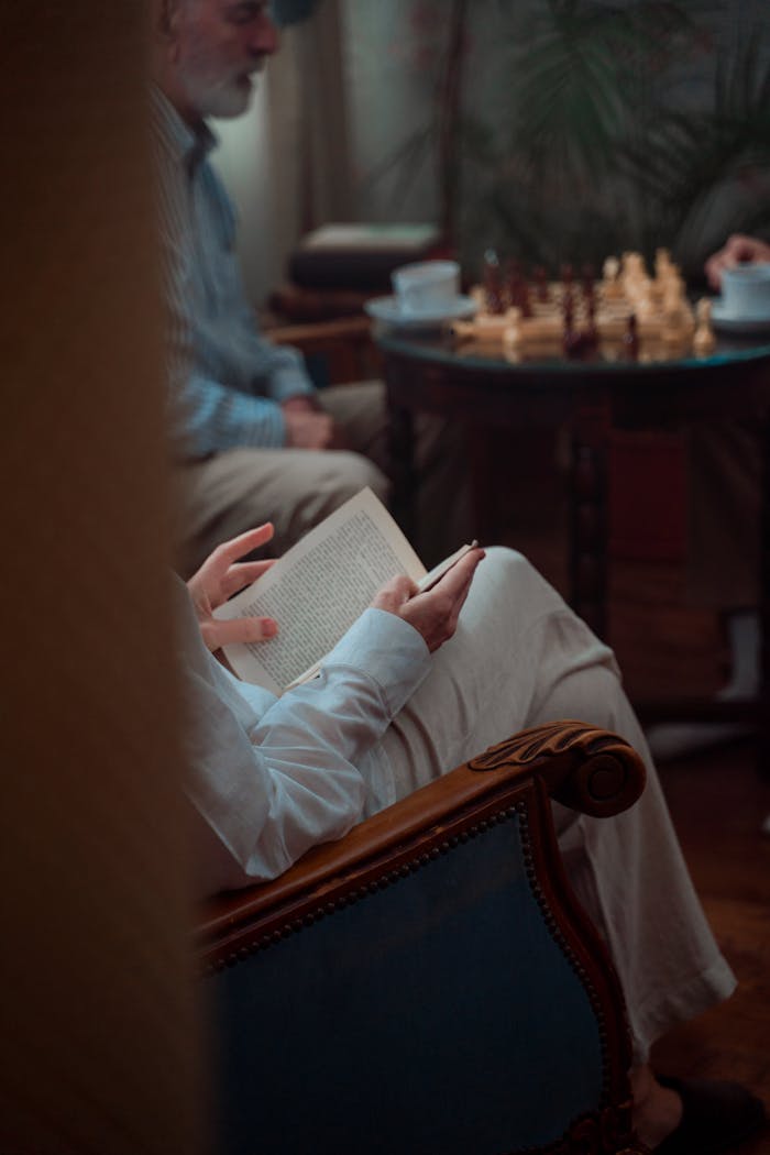 Elderly man reading a book indoors as others play chess on a table.